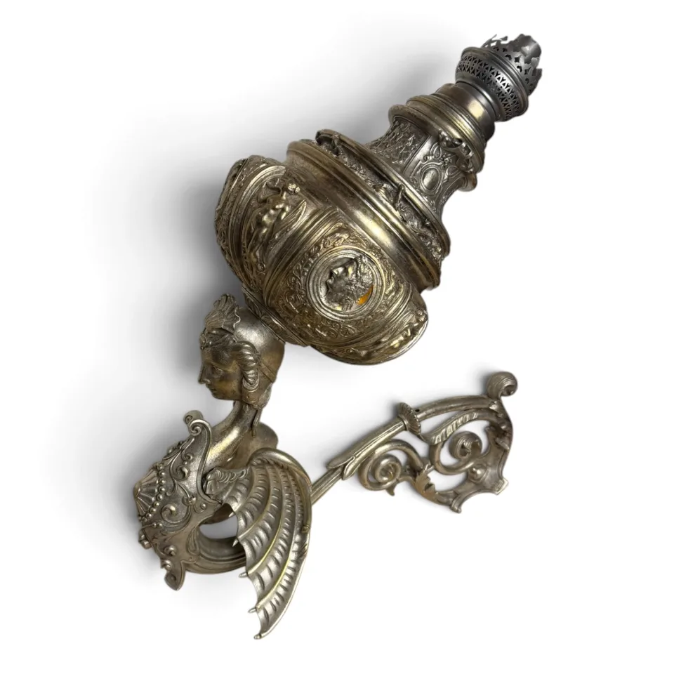 Lampa naftowa ścienna z metalu w kolorze srebra, z uskrzydloną figurą kobiecą podtrzymującą zbiornik, w stylu neorenesansowym.