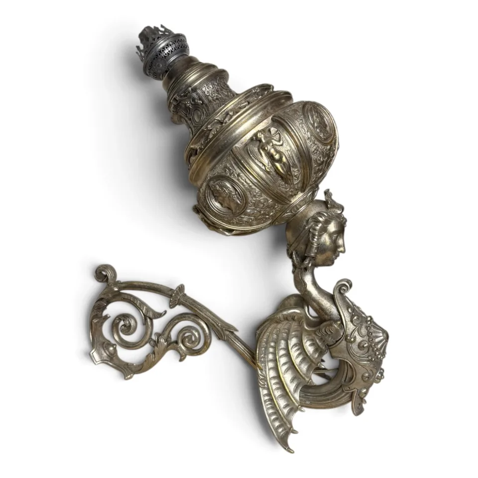Lampa naftowa ścienna z metalu w kolorze srebra, z uskrzydloną figurą kobiecą podtrzymującą zbiornik, w stylu neorenesansowym.